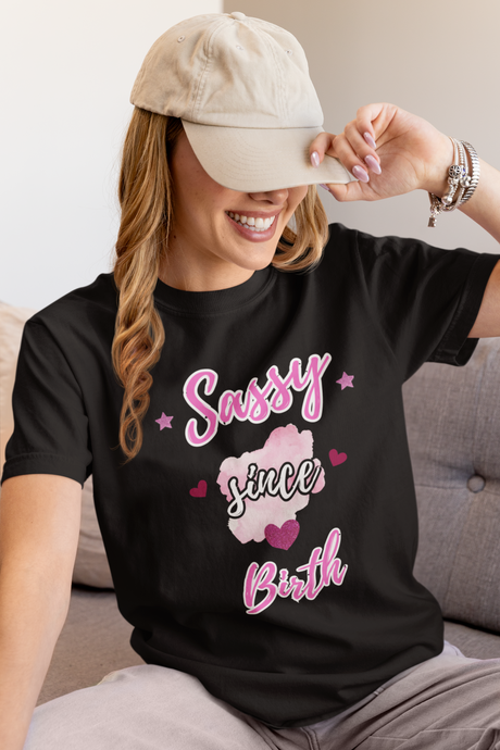 Woman wearing a black t-shirt with 'Sassy since birth' text and pink graphics, sitting on a couch.